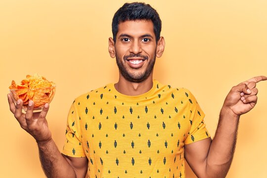 Young latin man holding nachos potato chips smiling happy pointing with hand and finger to the side