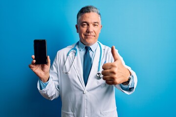 Middle age handsome grey-haired doctor man holding smartphone showing screen happy with big smile doing ok sign, thumb up with fingers, excellent sign