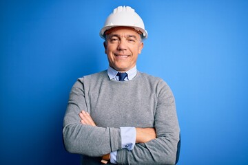 Middle age handsome grey-haired engineer man wearing safety helmet over blue background happy face smiling with crossed arms looking at the camera. Positive person.