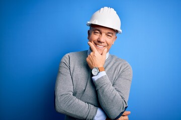 Middle age handsome grey-haired engineer man wearing safety helmet over blue background looking confident at the camera smiling with crossed arms and hand raised on chin. Thinking positive.