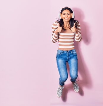 Young beautiful hispanic woman smiling happy. Jumping with smile on face listening to music using headphones doing ok sign with thums up over pink background
