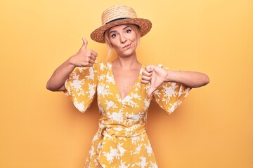 Beautiful blonde woman on vacation wearing summer hat and dress over yellow background Doing thumbs up and down, disagreement and agreement expression. Crazy conflict