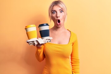 Young beautiful blonde woman holding cups of takeaway coffee over isolated yellow background scared and amazed with open mouth for surprise, disbelief face