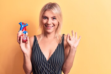 Young beautiful blonde woman asking for health care holding heart with veins and arteries doing ok sign with fingers, smiling friendly gesturing excellent symbol