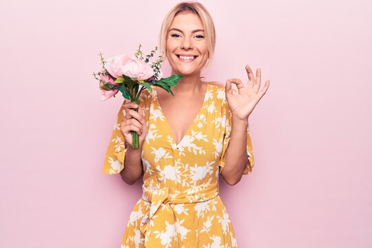 Young beautiful blonde woman holding bouquet of flowers over isolated pink background doing ok sign with fingers, smiling friendly gesturing excellent symbol