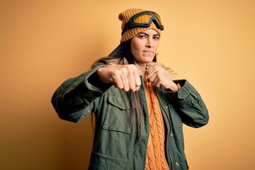 Young beautiful hispanic woman wearing ski glasses and coat for winter weather Punching fist to fight, aggressive and angry attack, threat and violence