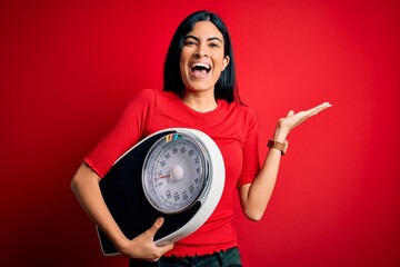 Young beautiful hispanic fitness woman holding scale for healthy weight over red background very...