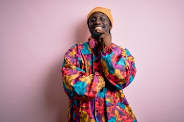 Young handsome african american man wearing colorful coat and cap over pink background looking confident at the camera with smile with crossed arms and hand raised on chin. Thinking positive.