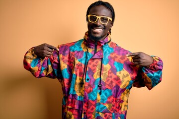 Handsome african american man wearing colorful coat and glasses over yellow background looking confident with smile on face, pointing oneself with fingers proud and happy.