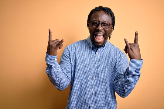 Young handsome african american man wearing shirt and glasses over yellow background shouting with crazy expression doing rock symbol with hands up. Music star. Heavy music concept.