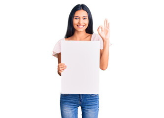 Young beautiful latin girl holding blank empty banner doing ok sign with fingers, smiling friendly gesturing excellent symbol