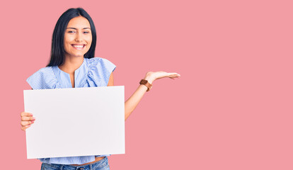 Young beautiful latin girl holding blank empty banner celebrating victory with happy smile and winner expression with raised hands