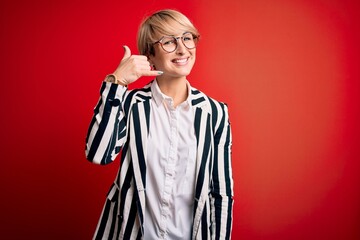 Blonde business woman with short hair wearing glasses and striped jacket over red background smiling doing phone gesture with hand and fingers like talking on the telephone. Communicating concepts.