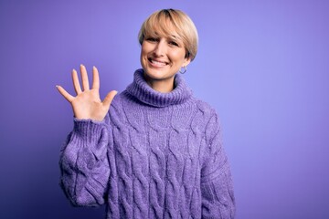 Young blonde woman with short hair wearing winter turtleneck sweater over purple background showing and pointing up with fingers number five while smiling confident and happy.