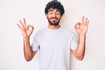 Handsome young man with curly hair and bear wearing casual tshirt relax and smiling with eyes closed doing meditation gesture with fingers. yoga concept.