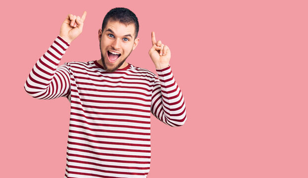 Young handsome man wearing striped sweater smiling amazed and surprised and pointing up with fingers and raised arms.
