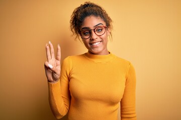 Young beautiful african american girl wearing sweater and glasses over yellow background showing and pointing up with fingers number three while smiling confident and happy.