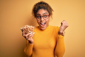 Young african american afro girl holding bowl with healthy peanuts over yellow background screaming proud and celebrating victory and success very excited, cheering emotion
