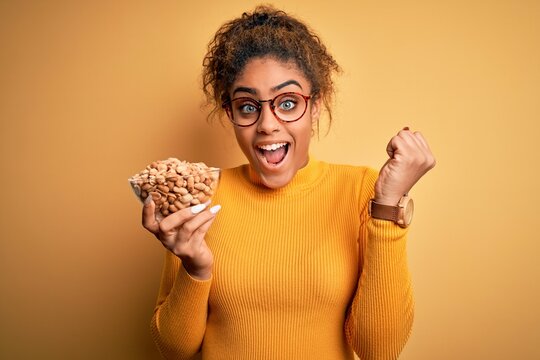 Young african american afro girl holding bowl with healthy peanuts over yellow background screaming proud and celebrating victory and success very excited, cheering emotion