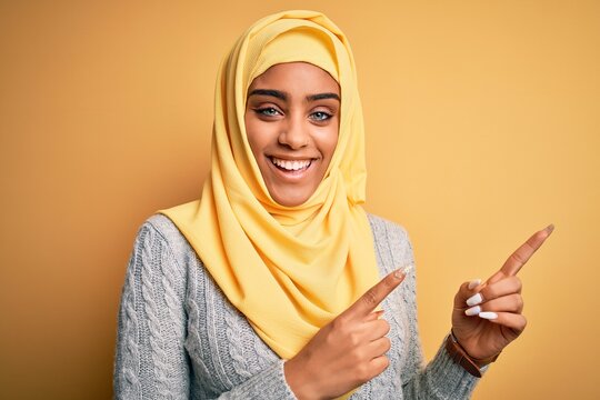 Young beautiful african american girl wearing muslim hijab over isolated yellow background smiling and looking at the camera pointing with two hands and fingers to the side.