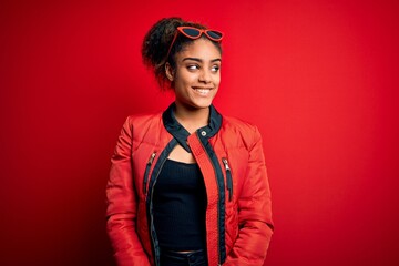 Beautiful african american girl wearing red jacket and sunglasses over isolated background looking away to side with smile on face, natural expression. Laughing confident.