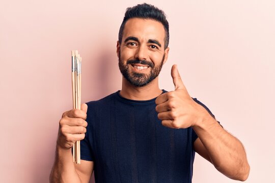 Young hispanic man holding paintbrushes smiling happy and positive, thumb up doing excellent and approval sign