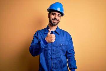 Mechanic man with beard wearing blue uniform and safety helmet over yellow background doing happy...