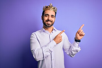 Young handsome man with beard wearing golden crown of king over purple background smiling and looking at the camera pointing with two hands and fingers to the side.