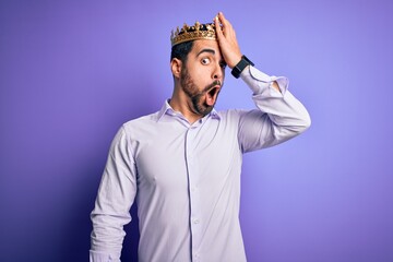 Young handsome man with beard wearing golden crown of king over purple background surprised with...
