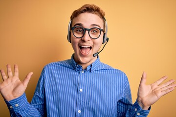 Young handsome redhead call center agent man wearing glasses working using headset celebrating crazy and amazed for success with arms raised and open eyes screaming excited. Winner concept