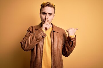 Young handsome redhead man wearing casual leather jacket over isolated yellow background asking to be quiet with finger on lips pointing with hand to the side. Silence and secret concept.