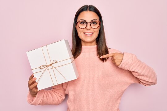 Young beautiful brunette woman holding birthday gift over isolated pink background pointing finger to one self smiling happy and proud