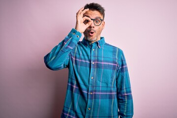 Young handsome man wearing casual shirt and glasses standing over isolated pink background doing ok gesture shocked with surprised face, eye looking through fingers. Unbelieving expression.
