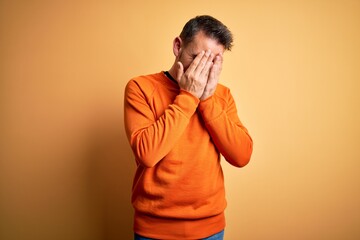 Young handsome man wearing orange casual sweater standing over isolated yellow background with sad expression covering face with hands while crying. Depression concept.