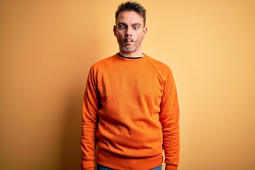 Young handsome man wearing orange casual sweater standing over isolated yellow background making fish face with lips, crazy and comical gesture. Funny expression.