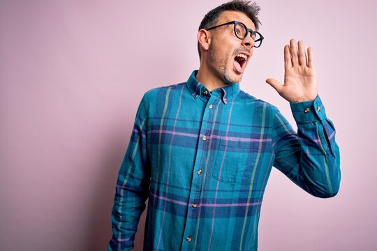 Young handsome man wearing casual shirt and glasses standing over isolated pink background shouting and screaming loud to side with hand on mouth. Communication concept.