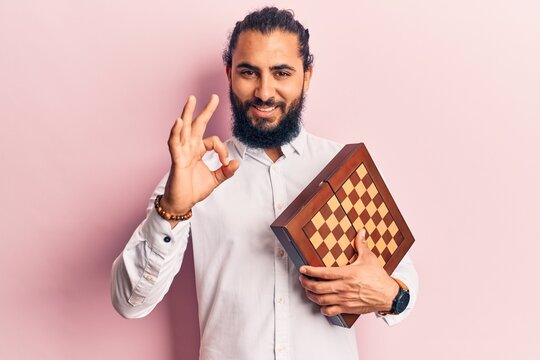 Young arab man holding chess doing ok sign with fingers, smiling friendly gesturing excellent symbol