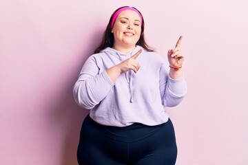 Young plus size woman wearing sportswear smiling and looking at the camera pointing with two hands and fingers to the side.