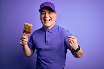 Young modern decorator painter man holding paint brush over purple background screaming proud and celebrating victory and success very excited, cheering emotion