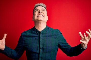 Young handsome modern man wearing elegant green shirt over red isolated background crazy and mad shouting and yelling with aggressive expression and arms raised. Frustration concept.