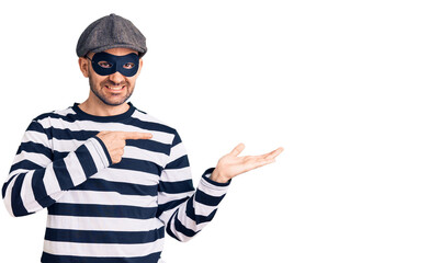 Young handsome man wearing burglar mask amazed and smiling to the camera while presenting with hand and pointing with finger.