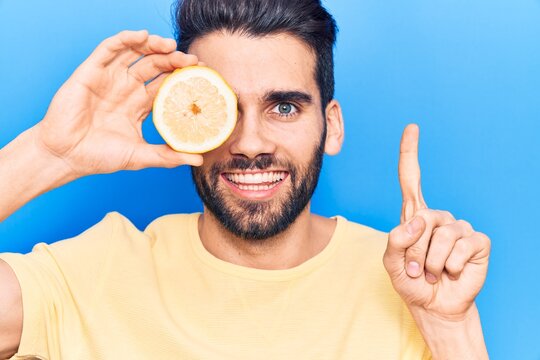 Young handsome man with beard holding slice of lemon over eye smiling with an idea or question pointing finger with happy face, number one - Powered by Adobe