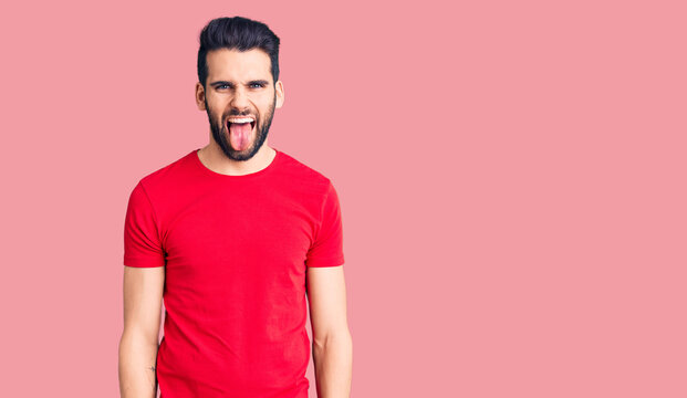 Young handsome man with beard wearing casual t-shirt sticking tongue out happy with funny expression. emotion concept.