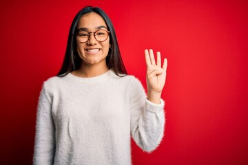 Young beautiful asian woman wearing casual sweater and glasses over red background showing and pointing up with fingers number four while smiling confident and happy.