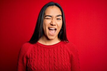 Young beautiful asian woman wearing casual sweater standing over isolated red background winking...