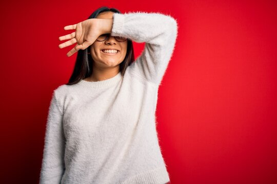 Young beautiful asian woman wearing casual sweater and glasses over red background covering eyes with arm smiling cheerful and funny. Blind concept.