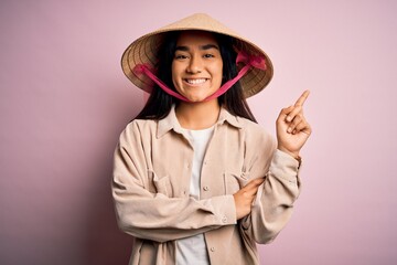 Young beautiful woman wearing traditional conical asian hat over isolated pink background with a big smile on face, pointing with hand and finger to the side looking at the camera.