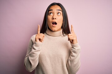 Young beautiful asian woman wearing casual turtleneck sweater over pink background amazed and surprised looking up and pointing with fingers and raised arms.