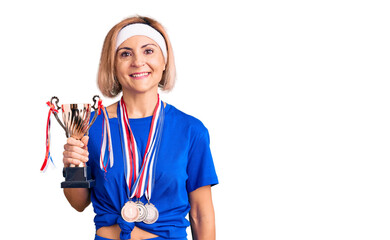 Young blonde woman holding champion trophy wearing medals looking positive and happy standing and smiling with a confident smile showing teeth