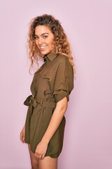 Young beautiful blonde woman with curly hair smiling happy and confident. Standing with smile on face wearing casual dress over isolated pink background